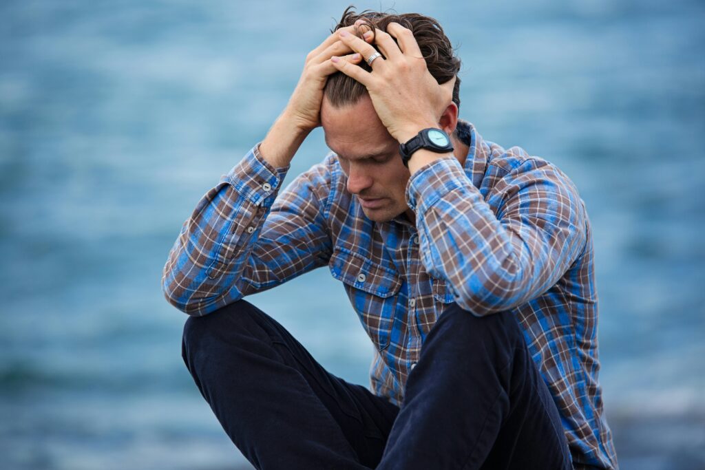Duelos: Al dolor por la pérdida, se suma el miedo a soltar. ¿qué viene después de que suelto eso que ya no está más? A man in a plaid shirt sits by the water looking distressed, symbolizing stress.